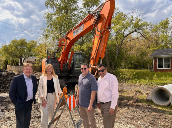 Township elected officials stand together on a construction site in front of a large orange excavator. They are dressed in business and business-casual attire and smiling at the camera. The ground is gravel with construction equipment and materials nearby, including a traffic barricade and a concrete pipe. Trees and residential homes are visible in the background under a partly cloudy sky.